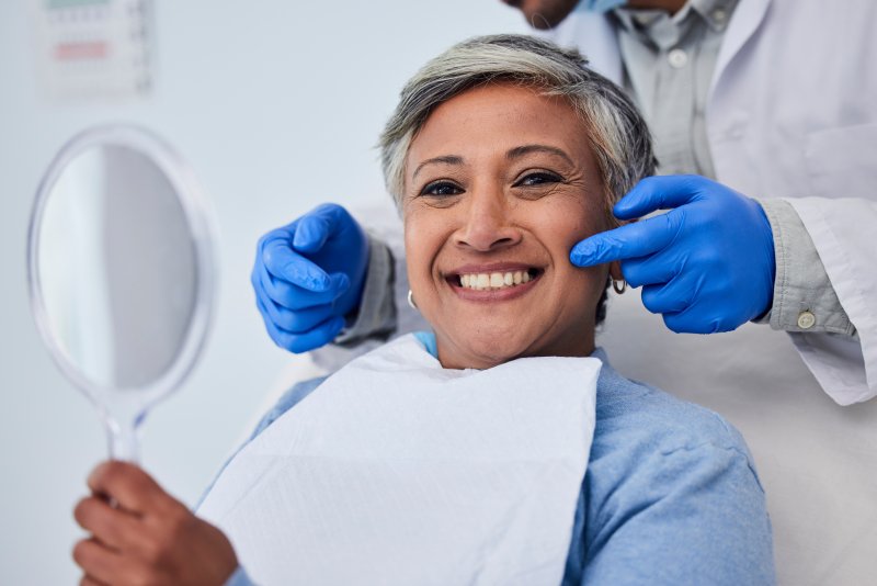 Older woman smiling with teeth at viewer while holding small mirror up to her face as dentist has two gloved hands on either sides of her cheeks